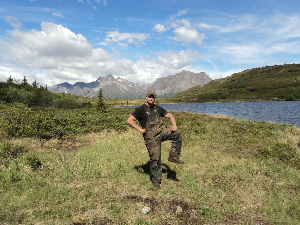 Man in dirty overalls stands with one leg up in a grassy field by a lake, with mountains in the background