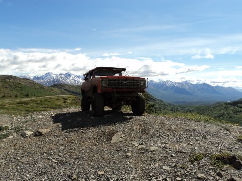 1990 Chevrolet Suburban - A lifted red SUV sits on a rocky incline, overlooking a vast mountain landscape under a partly cloudy sky.