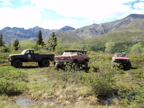1990 Chevrolet Suburban - Several off-road vehicles are parked in a field with mountains in the background.