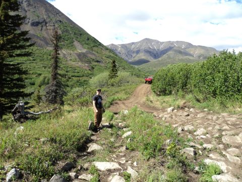 1990 Chevrolet Suburban - A red SUV navigates a rough dirt road through a scenic mountain landscape.