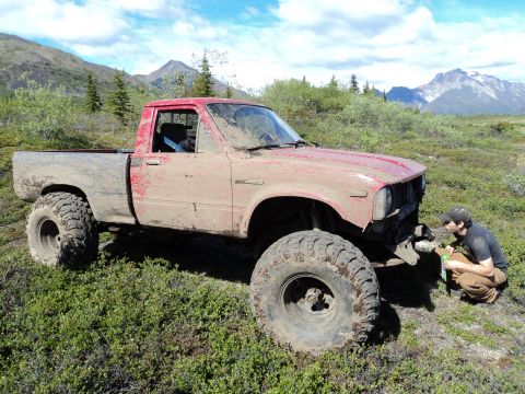 1990 Chevrolet Suburban - A heavily modified and mud-caked pickup truck sits in a grassy, mountainous landscape, its front bumper removed.