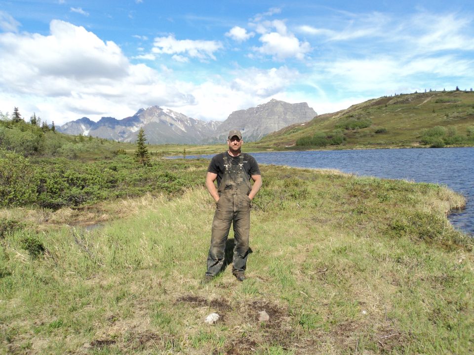 Man in dirty overalls stands in a grassy field near a lake with mountains behind him