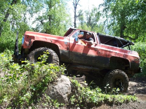 1990 Chevrolet Suburban - A modified red truck tackles a challenging off-road course amidst lush greenery.