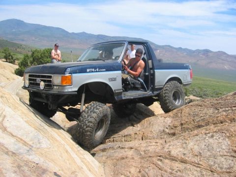 1989 Ford Bronco - This lifted pickup truck navigates a rocky trail under a clear blue sky.
