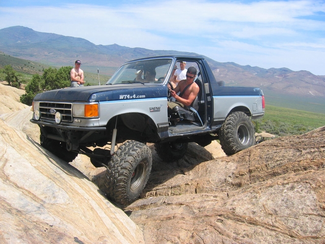 Modified pickup truck with large tires climbing rocks off-road. Driver and passenger visible