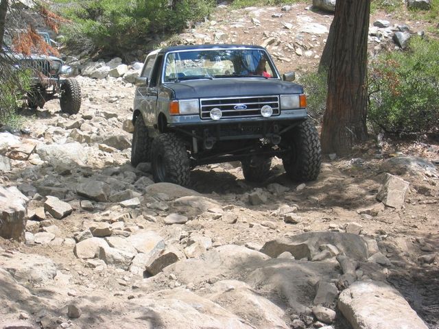 Lifted blue and silver Ford Bronco crawling a rocky forest trail on mud-terrain tires