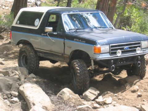 1989 Ford Bronco - This lifted Ford Bronco is shown tackling a challenging off-road course with large rocks and dirt.