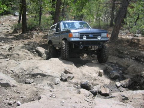 1989 Ford Bronco - A modified Ford Bronco tackles a challenging rocky trail in a wooded area.