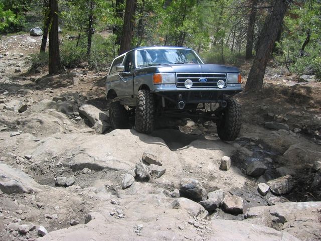 Topless blue Ford Bronco crawling down a rocky forest trail, front three-quarter view