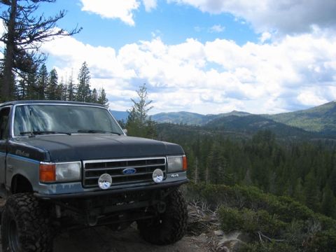 1989 Ford Bronco - A modified Ford truck is parked on a mountain trail with a scenic view of forested hills under a cloudy sky.