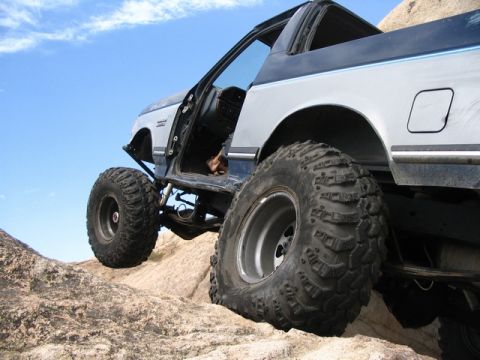 1989 Ford Bronco - A heavily modified SUV navigates rocky terrain under a clear blue sky.