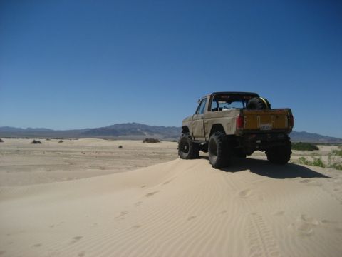 1989 Ford Bronco - This 1989 Ford Bronco is captured in a rear 3/4 view as it navigates a sandy terrain under a clear blue sky.