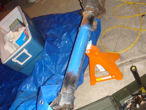 1992 Ford Bronco - A drive shaft, partially painted blue and showing rust, rests on a blue tarp with a cooler and jack stand nearby.