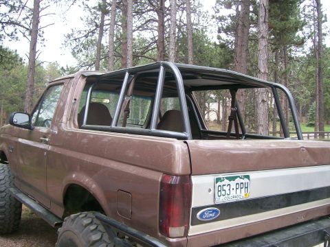 1993 Ford Bronco - Side-rear view of a lifted brown Ford Bronco with its hardtop removed, showing a black multi-hoop roll cage tied into the rear tub and a Co…
