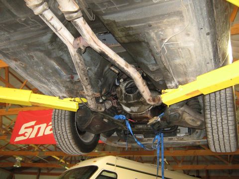 1985 Ford Mustang - The underside of a vehicle is shown while it is elevated on a car lift, revealing the exhaust system and transmission components.