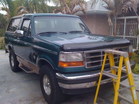 1996 Ford Bronco - A green 1996 Ford Bronco is viewed from a three-quarter front angle, parked in a driveway with palm trees and a house in the background.