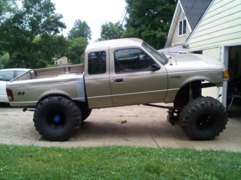 1997 Ford Ranger - Side view of a tan lifted Ford Ranger 4x4 extended cab pickup on big mud tires and steel wheels, parked in a driveway by a house.