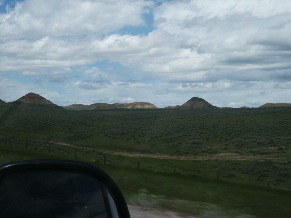 Black side mirror reflects a vast, green, rolling landscape under a cloudy sky