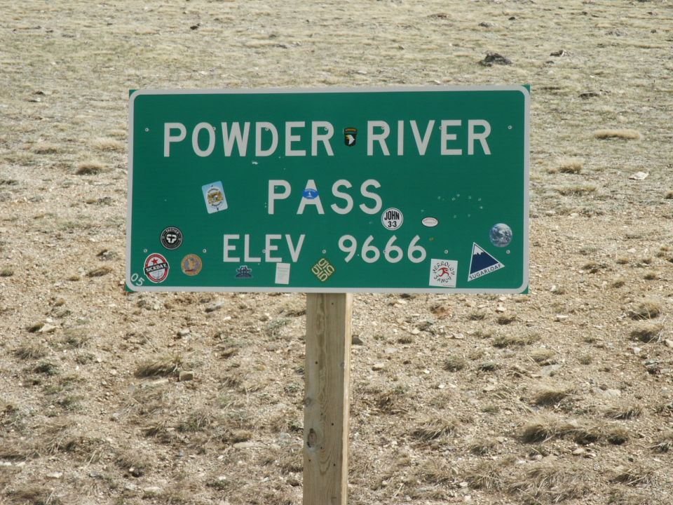 Green highway sign reading POWDER RIVER PASS ELEV 9666 covered in stickers