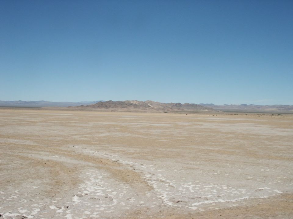 Wide desert landscape with mountains in the distance under a clear blue sky. Watermark reads SUPERMOTORS.NET