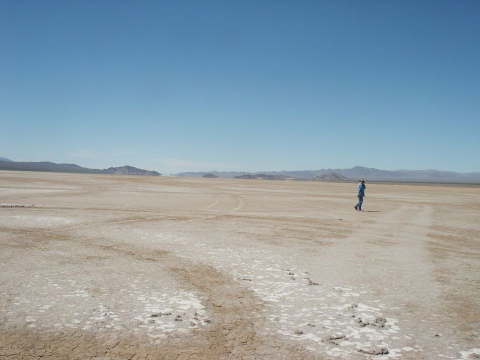 Person walking across a dry, cracked earth landscape under a clear blue sky. Mountains are visible in the distance