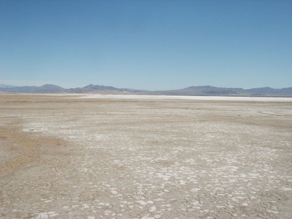 Wide shot of a dry, cracked salt flat with mountains in the distance under a clear blue sky. Supermotors logo in the corner