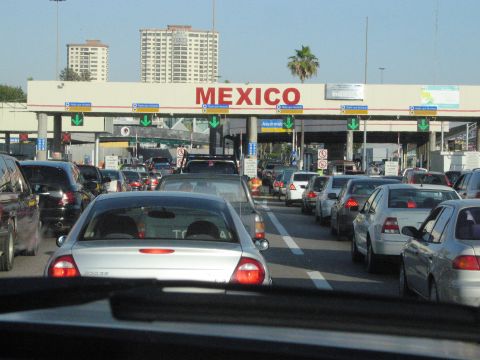 1986 Ford Bronco - Cars are backed up at a border crossing into Mexico, with large buildings visible in the background.