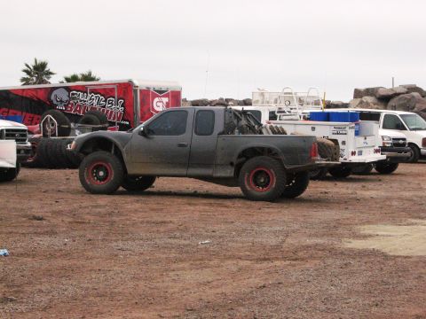 1986 Ford Bronco - A modified pickup truck with off-road tires and red rims is parked outdoors with other vehicles and trailers.