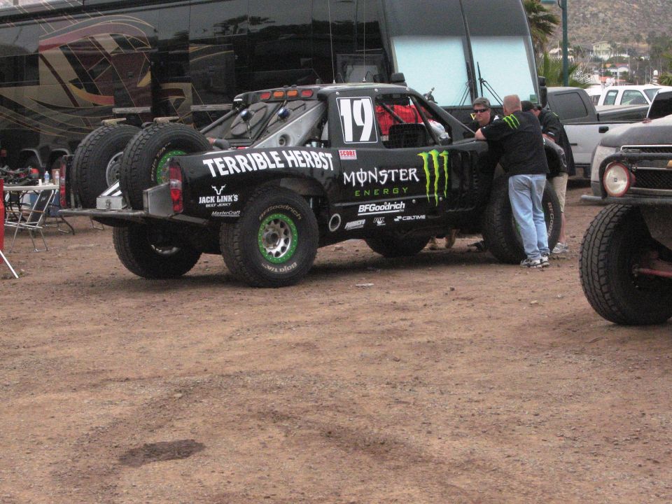 Monster Energy sponsored off-road truck with number 19 and 'TERRIBLE HERBST' text, parked near a bus and another vehicle