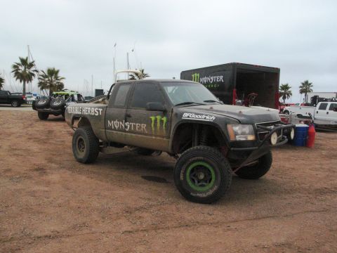 1986 Ford Bronco - A modified pickup truck with racing decals is parked on a dirt lot near boats and palm trees.
