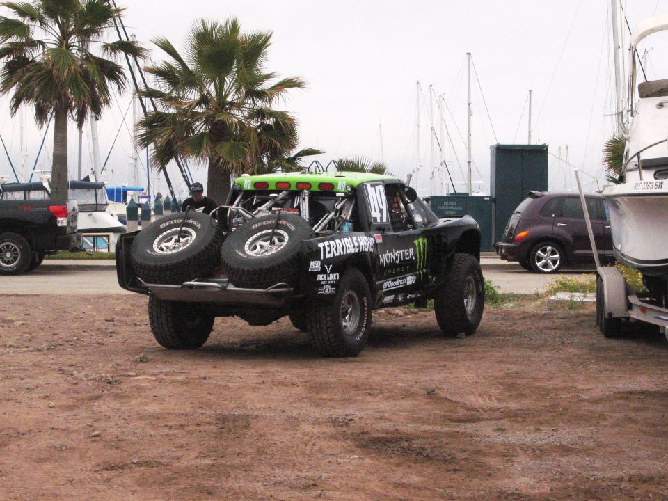 Off-road race truck with two spare tires mounted on the back, parked near boats and palm trees