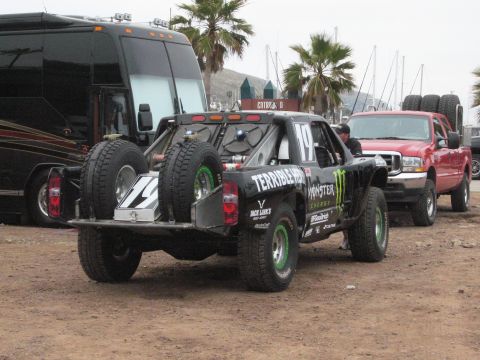 1986 Ford Bronco - A black off-road truck with racing decals is parked next to a red pickup truck and a bus.
