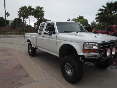 1986 Ford Bronco - Modified white Ford F-250 pickup truck parked curbside with palm trees in background.
