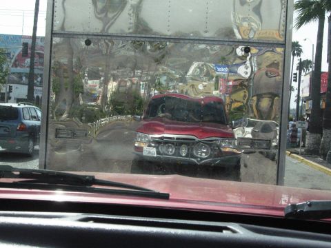 1986 Ford Bronco - A red truck with a custom front end is reflected in a shiny surface, showing a street scene with palm trees.