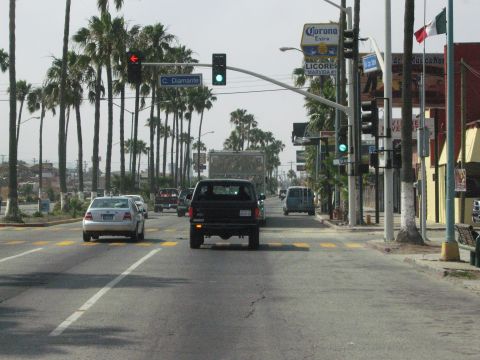 1986 Ford Bronco - A black SUV drives through an intersection lined with palm trees and businesses.