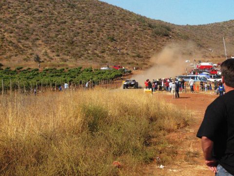 1986 Ford Bronco - An off-road race truck speeds down a dusty track lined with spectators and vineyards, during an event like the Baja 500.