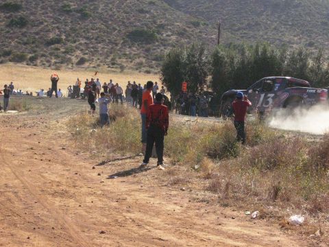 1986 Ford Bronco - A race truck speeds by spectators on a dusty off-road course.