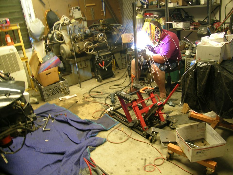 Man wearing a welding mask and purple shirt works on metal in a garage filled with tools and equipment