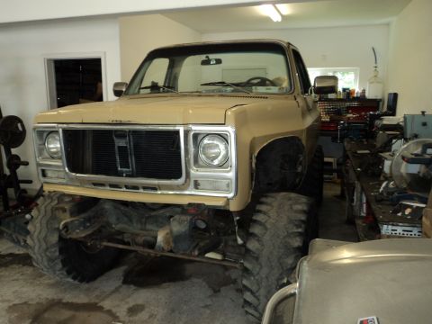 1990 Chevrolet Suburban - A tan square-body Chevrolet 4x4 truck sitting high on oversized mud tires inside a shop, shot from the front three-quarter angle. The hood…