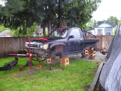 1990 Toyota Pickup/SR5 - A black pickup truck is elevated on jacks and blocks, with its suspension disassembled in a grassy yard.