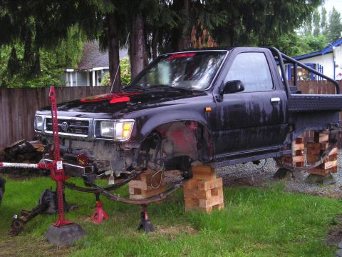 1990 Toyota Pickup/SR5 - A black Toyota pickup truck is elevated on wooden blocks and jack stands, undergoing front-end repairs.