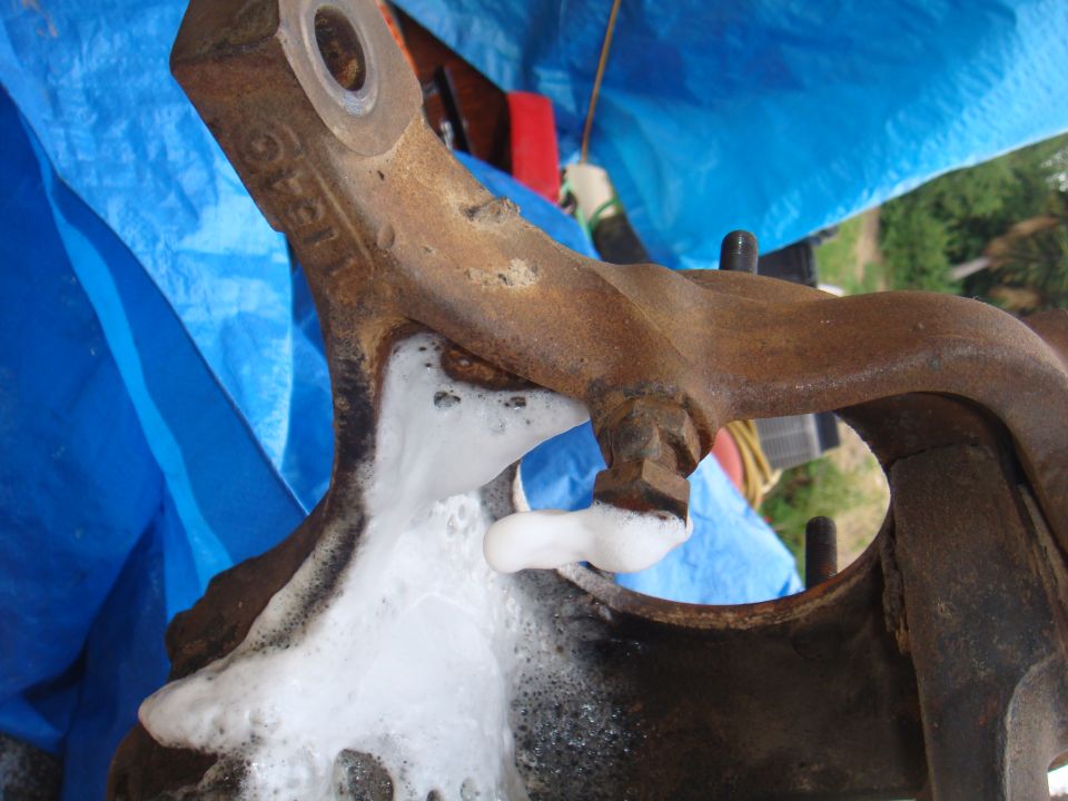 Rusty metal suspension component covered in soap suds, with a blue tarp in the background