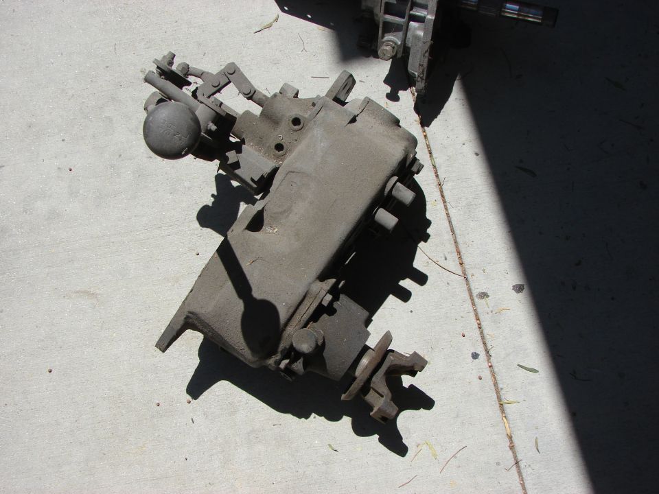 Gray metal transfer case with levers and a round knob, lying on concrete