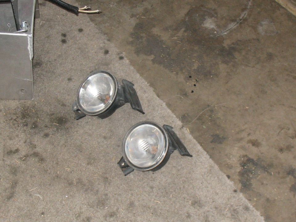 Pair of removed round clear-lens fog lights with black brackets on a garage floor