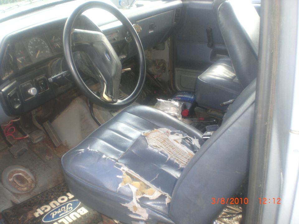 Interior shot of a truck's dashboard and ripped driver's seat. Floor mat says "Ford Tough Built"