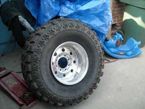 1985 Ford Bronco - A large off-road tire and chrome wheel assembly is shown, with a red jack and a blue toy dog nearby.