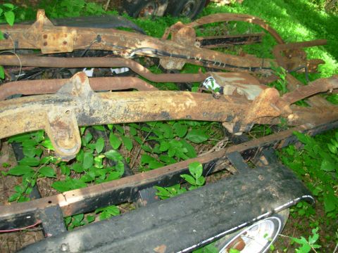 1976 Ford Bronco - Discarded vehicle frames and parts are piled up outdoors, covered in rust and overgrown with weeds.