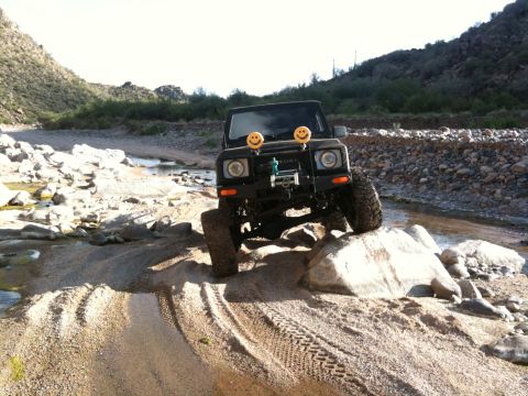 1987 Suzuki Samurai - A black 1987 Suzuki Samurai navigates a rocky stream bed, equipped with smiley face headlights and a front winch.