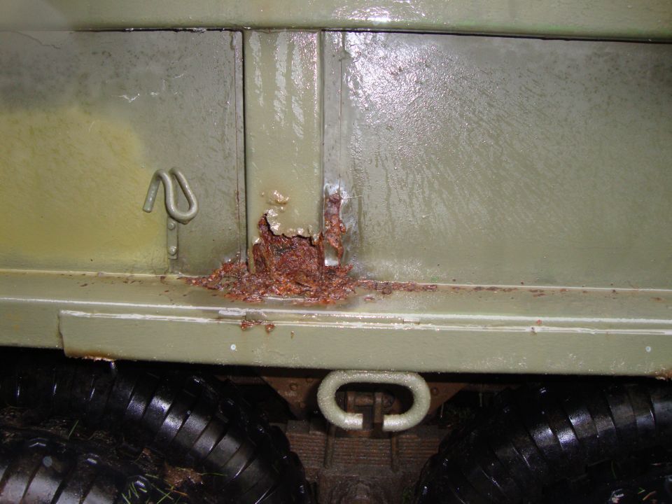 Rust blistering through olive drab paint on a military truck bed seam above dual tires