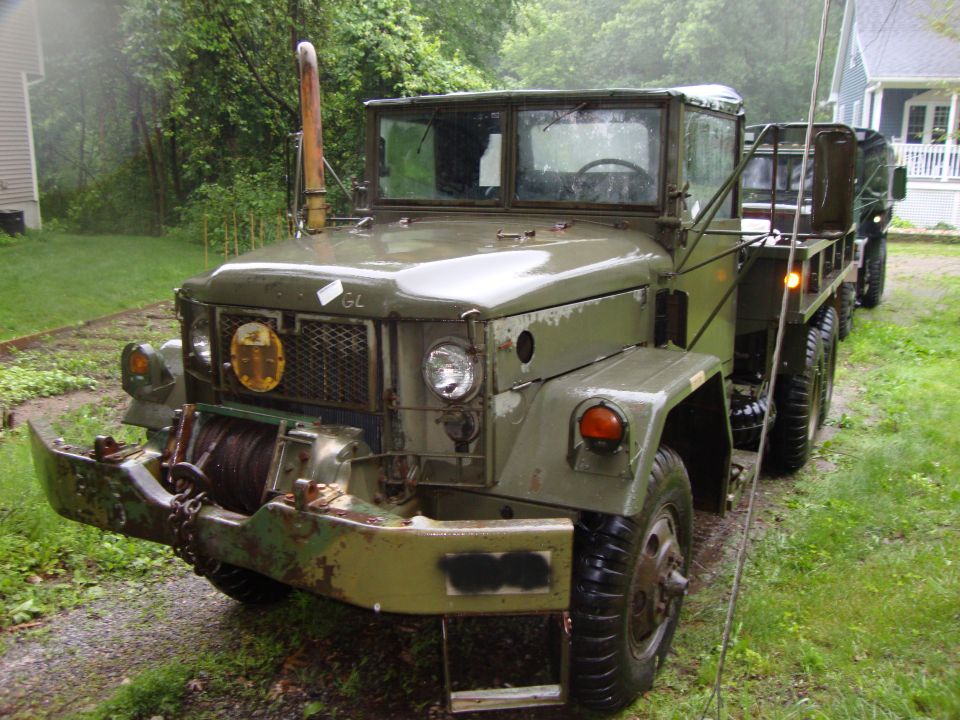 Olive drab military 6x6 cargo truck with front winch and exhaust stack on a wet driveway